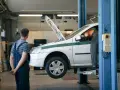 Dans un garage, un mécanicien en salopette bleue observe un autre mécanicien en orange qui travaille sous le capot d'une voiture blanche sur un pont élévateur. C'est essentiel pour entretenir sa voiture.