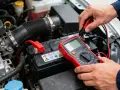 Main d'un mécanicien testant la batterie voiture sous le capot avec un multimètre rouge.