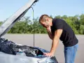 Homme désemparé devant le moteur de sa voiture, capot ouvert. Il semble avoir une panne batterie.