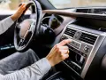 Main d'une femme ajustant la radio de la voiture, le volant à portée de main. La climatisation de la voiture est visible sur le tableau de bord.