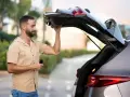 Un homme essaie de maintenir ouvert le coffre de sa voiture qui ne tient plus. Il tient une télécommande dans sa main.