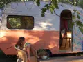 Une jeune femme lit, assise sur une chaise, devant une caravane vintage colorée, décorée de guirlandes lumineuses. La restauration de cette caravane a créé un espace cosy et charmant.