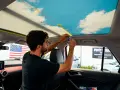 Un homme applique un adhésif pour recoller le ciel de toit d'une voiture, orné d'un motif de ciel bleu et de nuages.