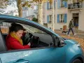 Femme frustrée tentant de démarrer sa voiture, un problème électrique voiture probable. En arrière-plan, un homme promène son chien.