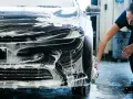 Une femme lave une voiture noire recouverte de mousse. Elle utilise une éponge pour nettoyer le pare-chocs, une étape essentielle pour savoir comment faire briller sa voiture.