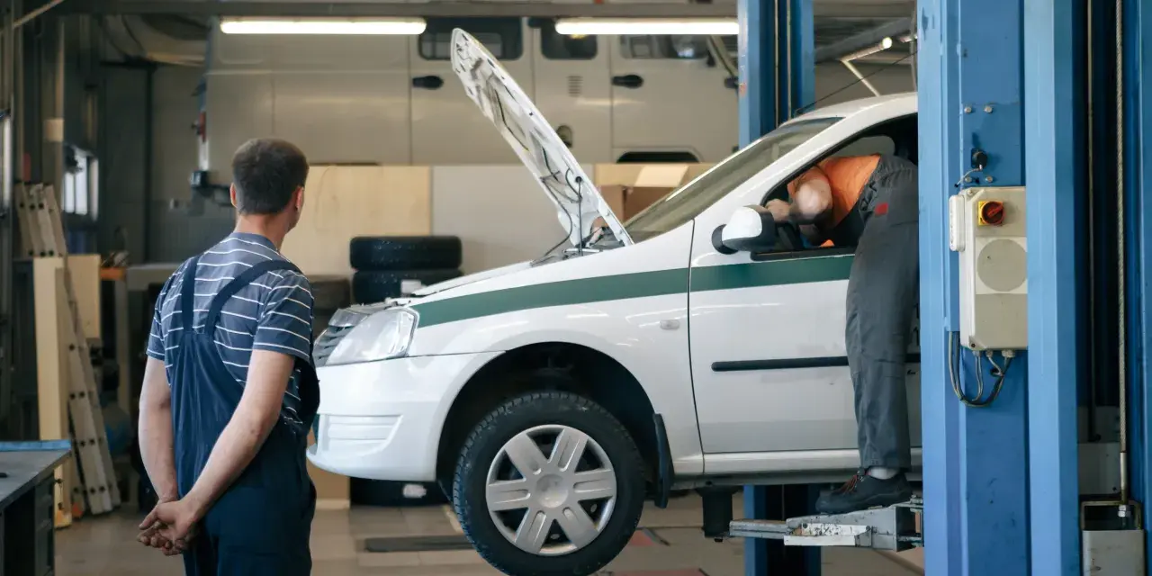Dans un garage, un mécanicien en salopette bleue observe un autre mécanicien en orange qui travaille sous le capot d'une voiture blanche sur un pont élévateur. C'est essentiel pour entretenir sa voiture.