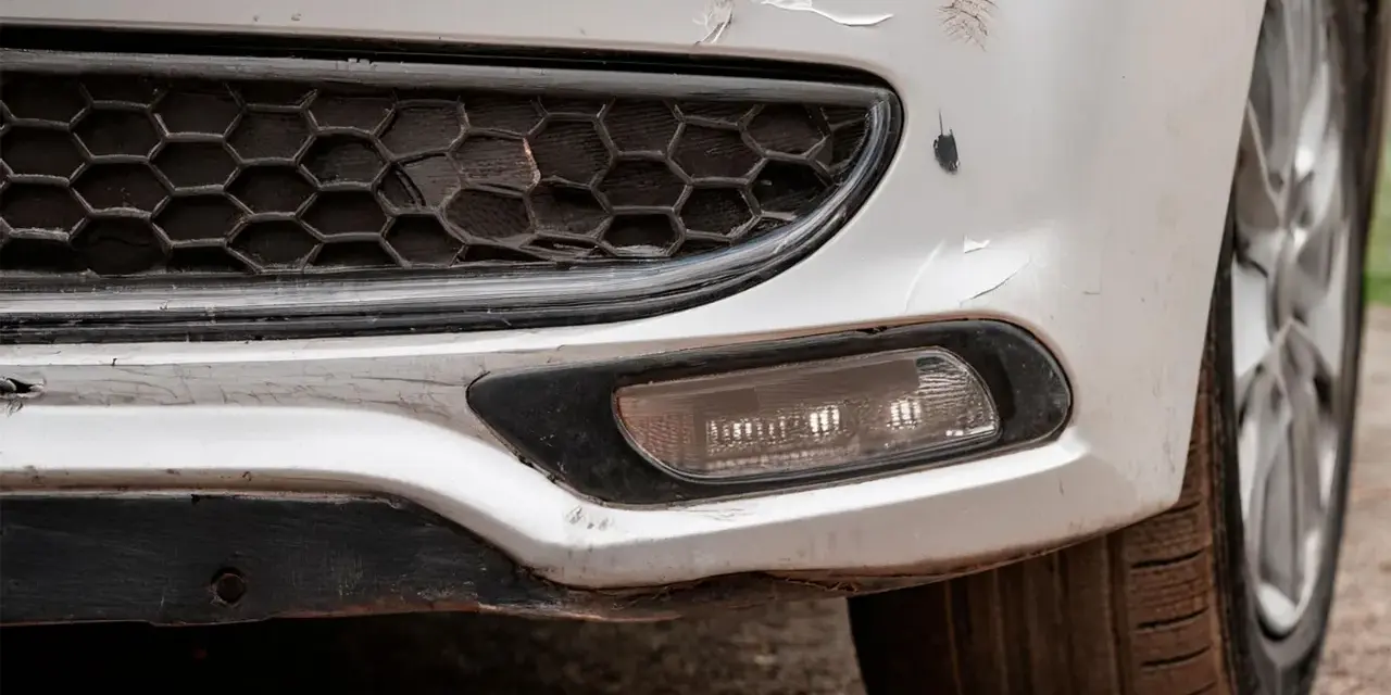 Pare-chocs avant d'une voiture blanche avec des éraflures et des éclats de peinture. Une méthode pour redresser carrosserie eau chaude pourrait aider à réparer ces dommages.