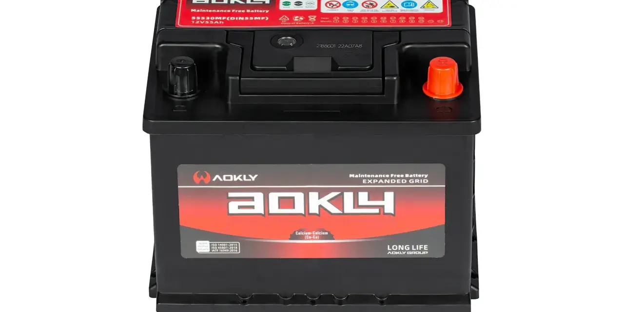 Une batterie AOKLY noire, 12V 55Ah, avec des étiquettes rouges et blanches indiquant "Maintenance Free Battery" et des symboles d'attention.