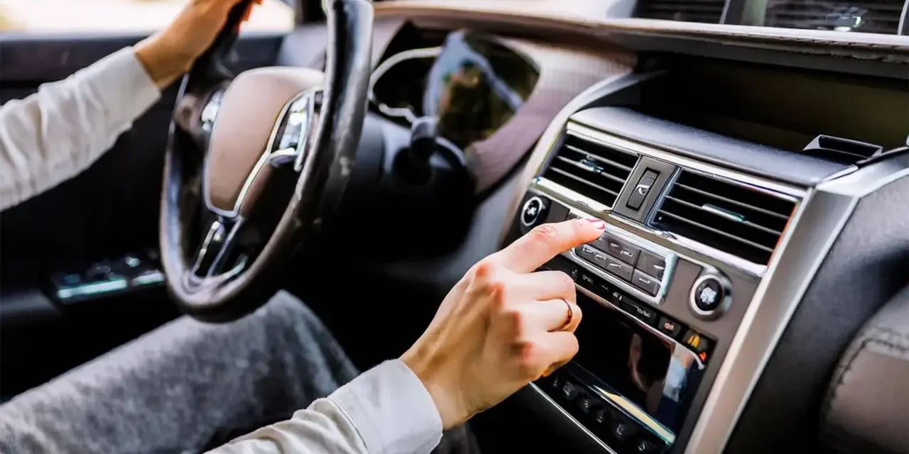 Main d'une femme ajustant la radio de la voiture, le volant à portée de main. La climatisation de la voiture est visible sur le tableau de bord.