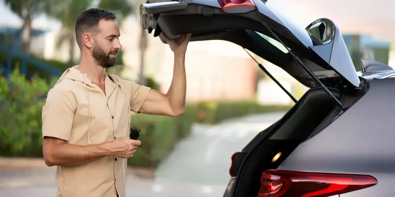 Un homme essaie de maintenir ouvert le coffre de sa voiture qui ne tient plus. Il tient une télécommande dans sa main.