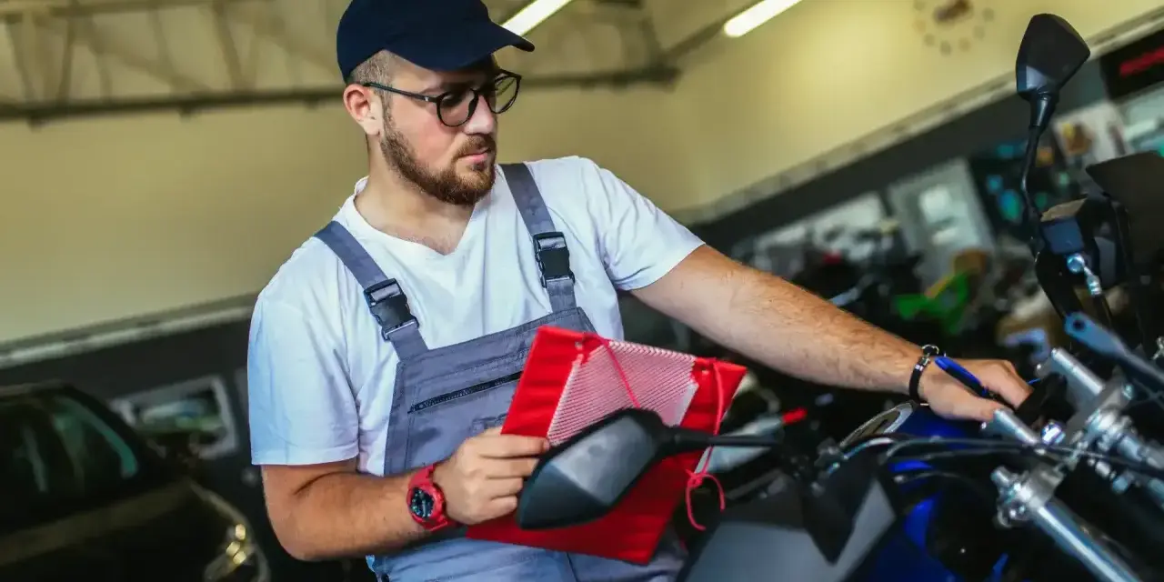 Un mécanicien en salopette grise vérifie un tableau rouge sur une moto bleue, assurant le pollution contrôle technique.
