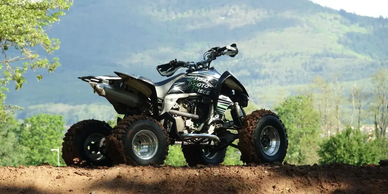 Quad tout-terrain, prêt pour son contrôle technique, avec des pneus boueux et un paysage montagneux en arrière-plan.