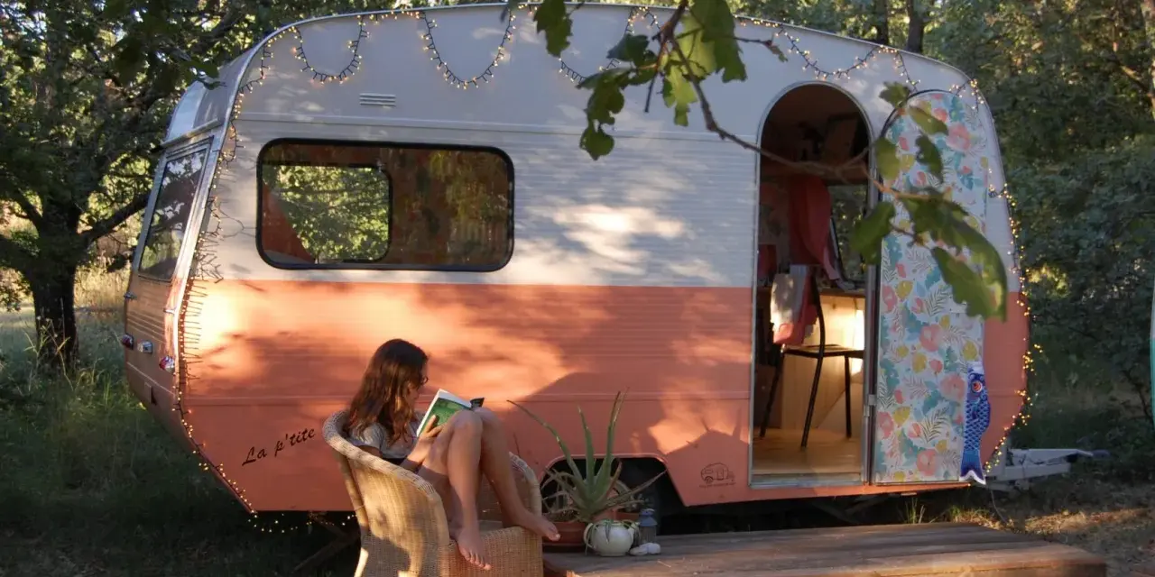 Une jeune femme lit, assise sur une chaise, devant une caravane vintage colorée, décorée de guirlandes lumineuses. La restauration de cette caravane a créé un espace cosy et charmant.