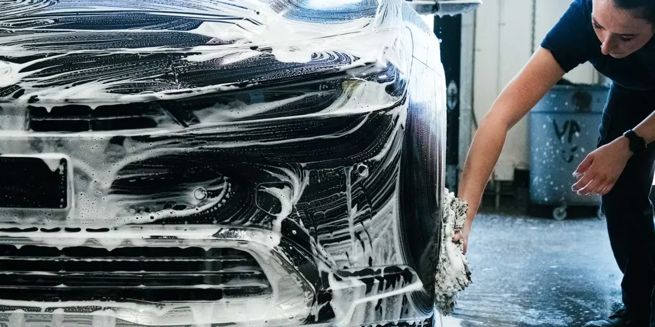 Une femme lave une voiture noire recouverte de mousse. Elle utilise une éponge pour nettoyer le pare-chocs, une étape essentielle pour savoir comment faire briller sa voiture.