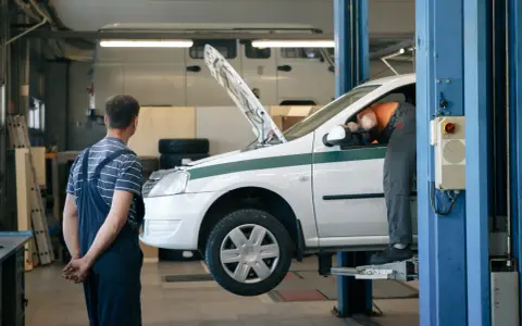 Dans un garage, un mécanicien en salopette bleue observe un autre mécanicien en orange qui travaille sous le capot d'une voiture blanche sur un pont élévateur. C'est essentiel pour entretenir sa voiture.