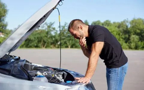 Homme désemparé devant le moteur de sa voiture, capot ouvert. Il semble avoir une panne batterie.