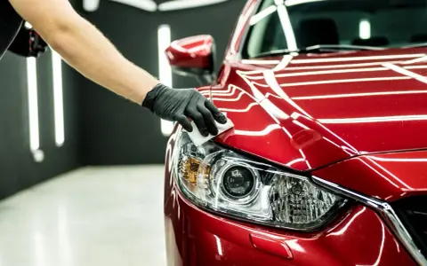 Main d'une personne gantée appliquant un produit sur le phare d'une voiture rouge. Une astuce de grand-mère pour faire briller une voiture.