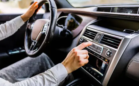 Main d'une femme ajustant la radio de la voiture, le volant à portée de main. La climatisation de la voiture est visible sur le tableau de bord.