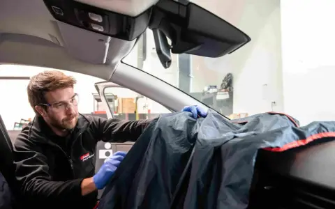 Un technicien en lunettes de protection utilise une housse pour protéger le tableau de bord lors d'une intervention sur la commande de désembuage.