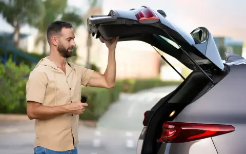 Un homme essaie de maintenir ouvert le coffre de sa voiture qui ne tient plus. Il tient une télécommande dans sa main.