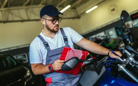 Un mécanicien en salopette grise vérifie un tableau rouge sur une moto bleue, assurant le pollution contrôle technique.