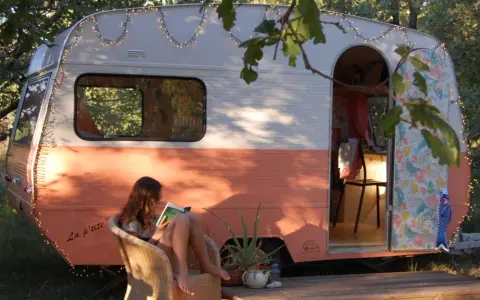 Une jeune femme lit, assise sur une chaise, devant une caravane vintage colorée, décorée de guirlandes lumineuses. La restauration de cette caravane a créé un espace cosy et charmant.