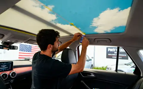 Un homme applique un adhésif pour recoller le ciel de toit d'une voiture, orné d'un motif de ciel bleu et de nuages.