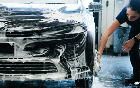 Une femme lave une voiture noire recouverte de mousse. Elle utilise une éponge pour nettoyer le pare-chocs, une étape essentielle pour savoir comment faire briller sa voiture.