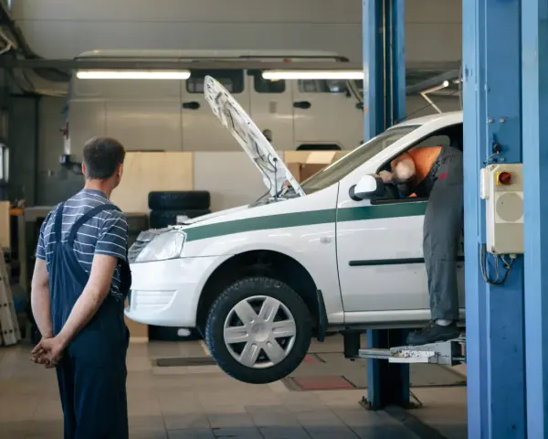 Dans un garage, un mécanicien en salopette bleue observe un autre mécanicien en orange qui travaille sous le capot d'une voiture blanche sur un pont élévateur. C'est essentiel pour entretenir sa voiture.