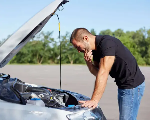 Homme désemparé devant le moteur de sa voiture, capot ouvert. Il semble avoir une panne batterie.