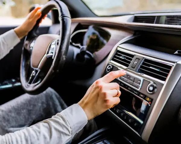 Main d'une femme ajustant la radio de la voiture, le volant à portée de main. La climatisation de la voiture est visible sur le tableau de bord.