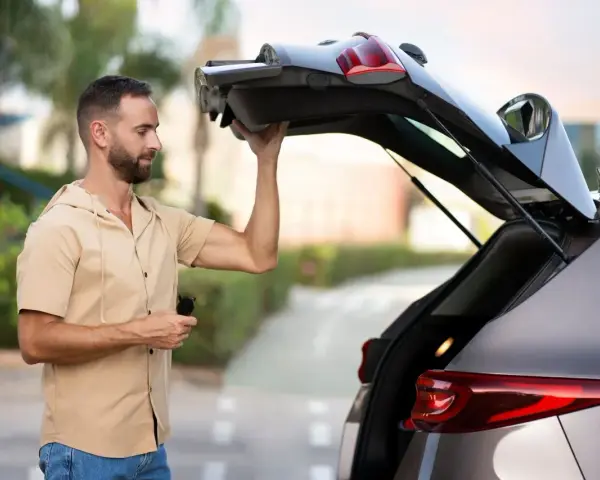 Un homme essaie de maintenir ouvert le coffre de sa voiture qui ne tient plus. Il tient une télécommande dans sa main.