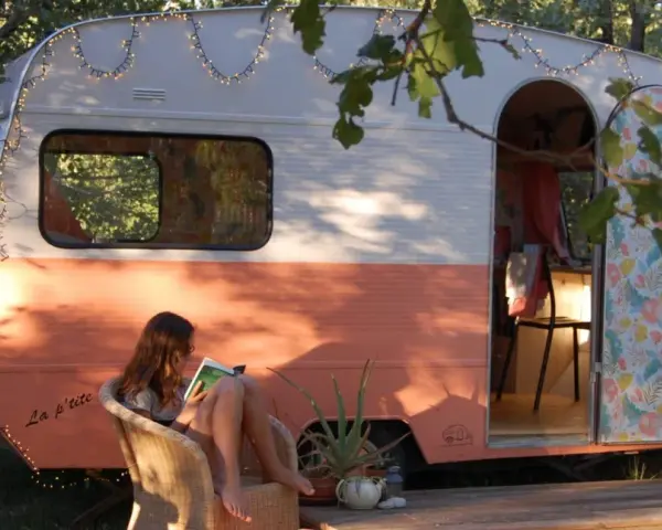 Une jeune femme lit, assise sur une chaise, devant une caravane vintage colorée, décorée de guirlandes lumineuses. La restauration de cette caravane a créé un espace cosy et charmant.