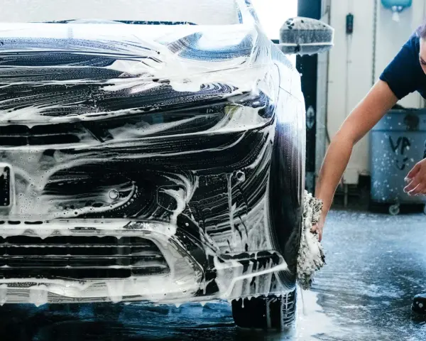 Une femme lave une voiture noire recouverte de mousse. Elle utilise une éponge pour nettoyer le pare-chocs, une étape essentielle pour savoir comment faire briller sa voiture.