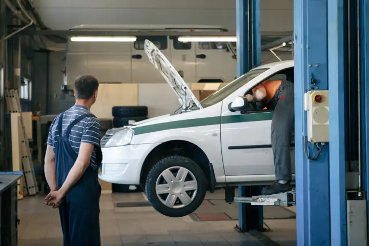 Dans un garage, un mécanicien en salopette bleue observe un autre mécanicien en orange qui travaille sous le capot d'une voiture blanche sur un pont élévateur. C'est essentiel pour entretenir sa voiture.