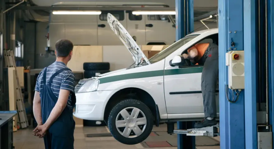 Dans un garage, un mécanicien en salopette bleue observe un autre mécanicien en orange qui travaille sous le capot d'une voiture blanche sur un pont élévateur. C'est essentiel pour entretenir sa voiture.