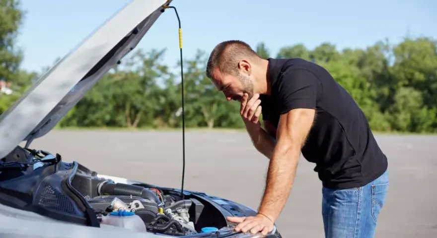 Homme désemparé devant le moteur de sa voiture, capot ouvert. Il semble avoir une panne batterie.