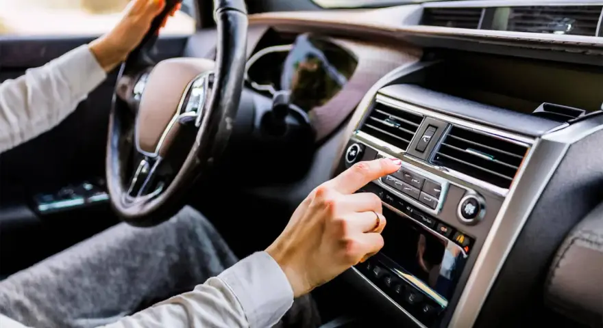 Main d'une femme ajustant la radio de la voiture, le volant à portée de main. La climatisation de la voiture est visible sur le tableau de bord.