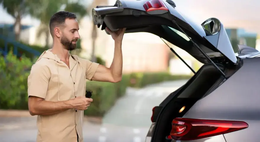 Un homme essaie de maintenir ouvert le coffre de sa voiture qui ne tient plus. Il tient une télécommande dans sa main.