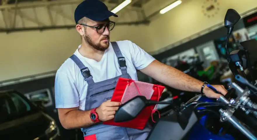 Un mécanicien en salopette grise vérifie un tableau rouge sur une moto bleue, assurant le pollution contrôle technique.