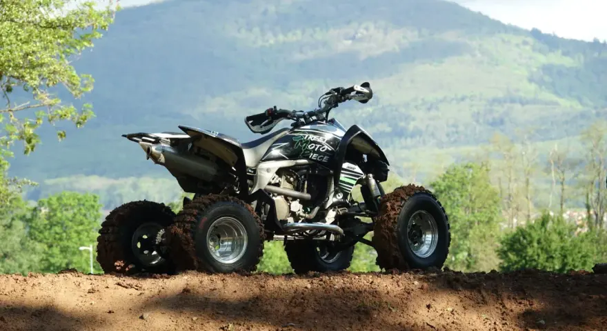 Quad tout-terrain, prêt pour son contrôle technique, avec des pneus boueux et un paysage montagneux en arrière-plan.
