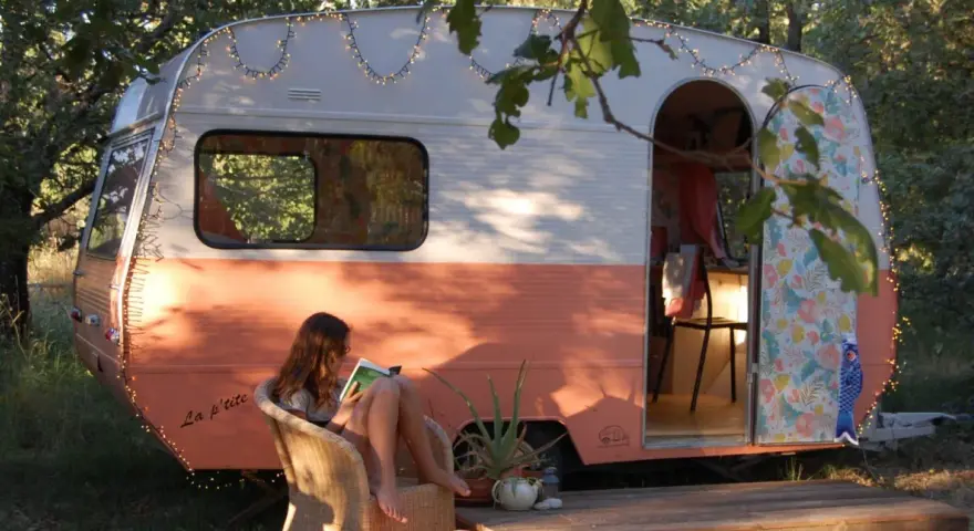 Une jeune femme lit, assise sur une chaise, devant une caravane vintage colorée, décorée de guirlandes lumineuses. La restauration de cette caravane a créé un espace cosy et charmant.