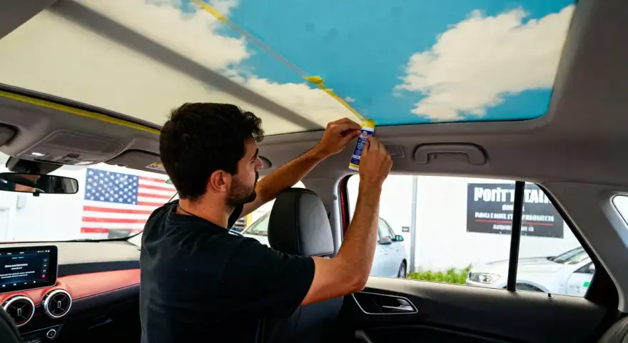 Un homme applique un adhésif pour recoller le ciel de toit d'une voiture, orné d'un motif de ciel bleu et de nuages.