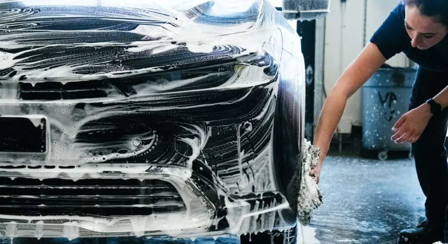 Une femme lave une voiture noire recouverte de mousse. Elle utilise une éponge pour nettoyer le pare-chocs, une étape essentielle pour savoir comment faire briller sa voiture.