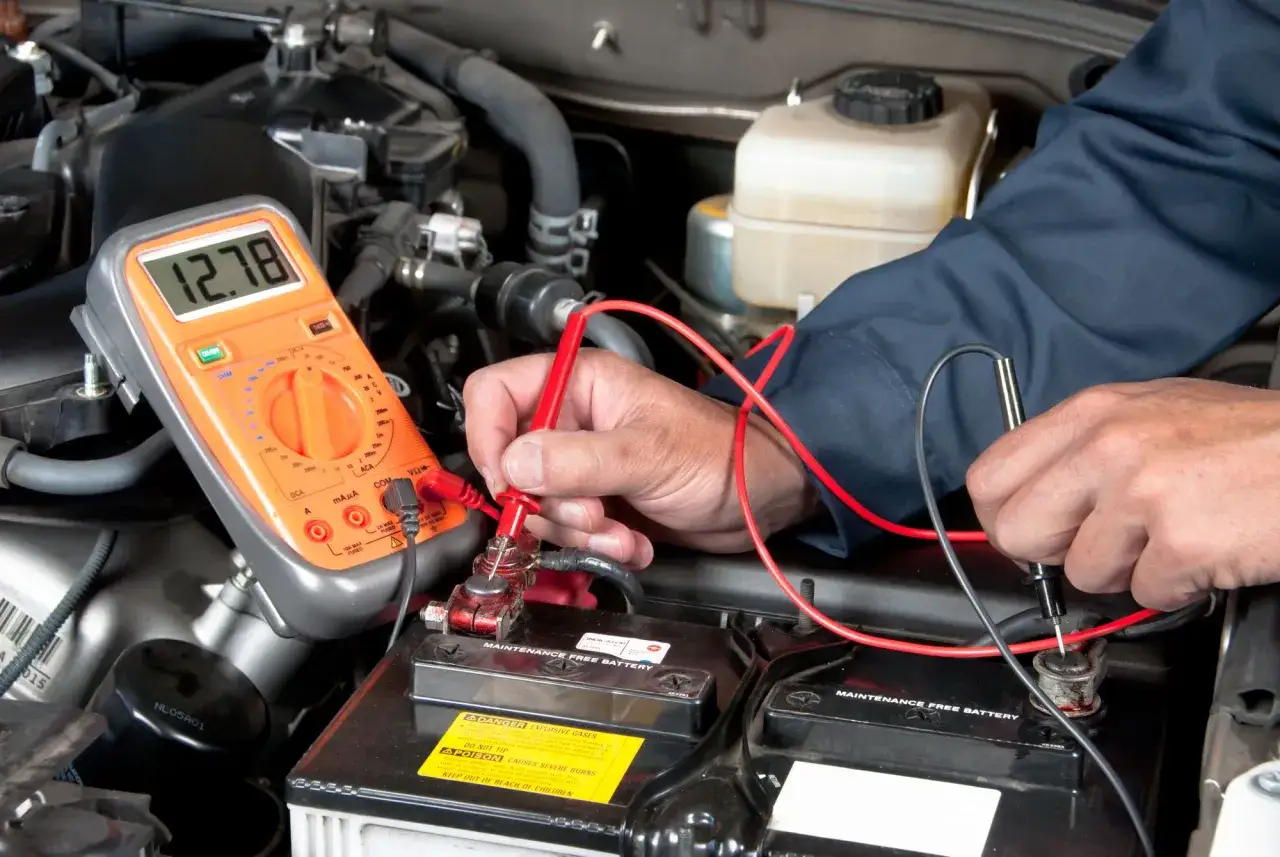 Un mécanicien vérifie la tension de la batterie voiture sous le capot avec un multimètre affichant 12.78V.
