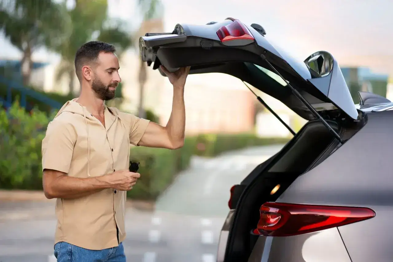 Un homme essaie de maintenir ouvert le coffre de sa voiture qui ne tient plus. Il tient une t&eacute;l&eacute;commande dans sa main.