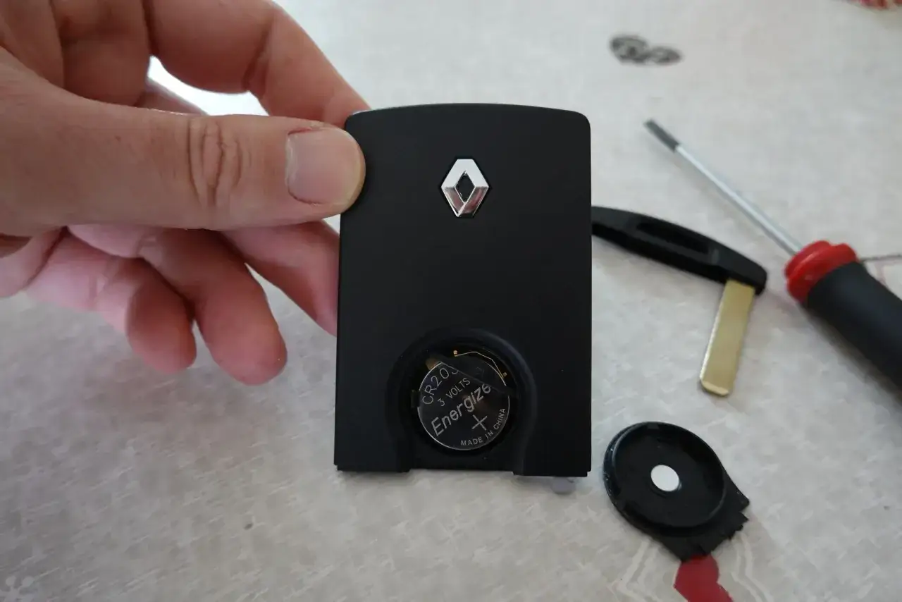 Main d'une personne tenant une carte Renault noire, pile CR2032 visible. Outil à proximité pour le changement de pile.
