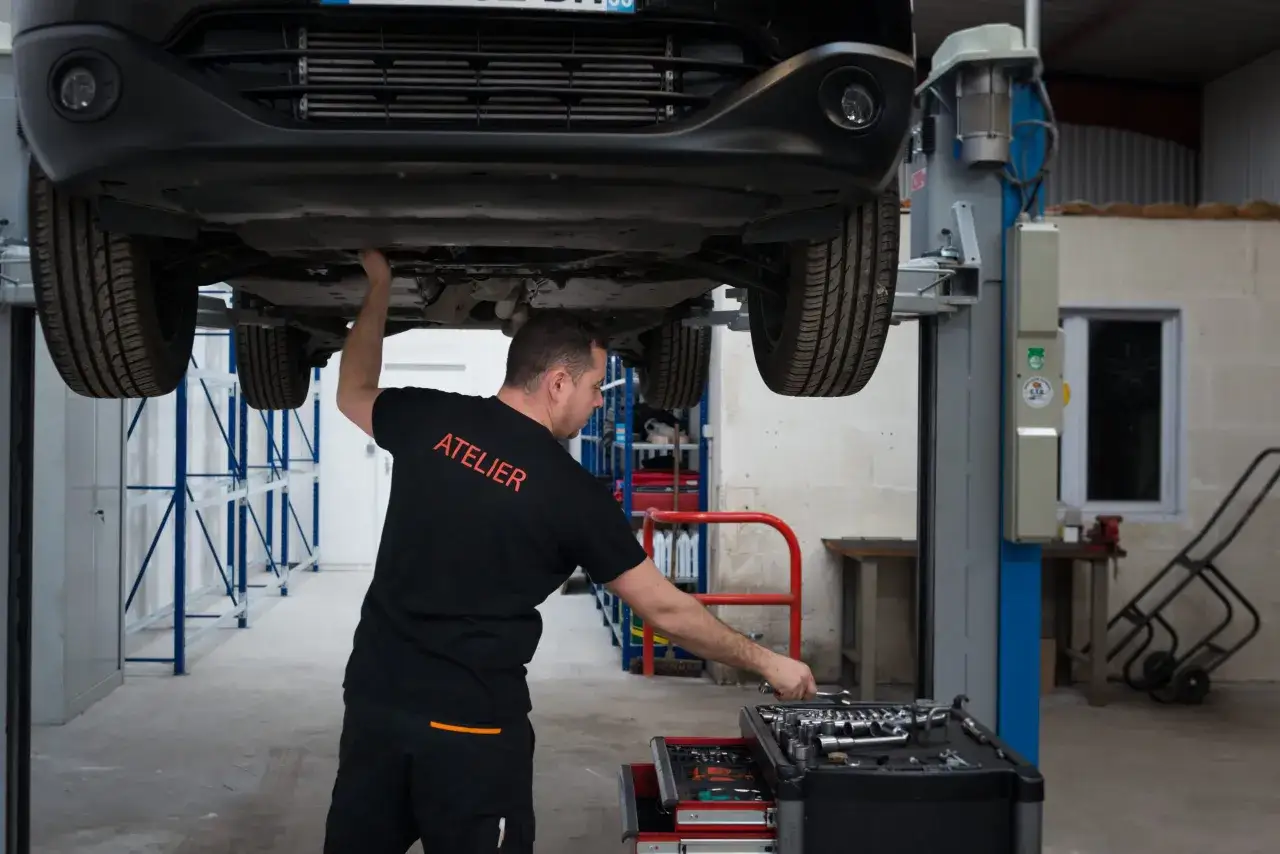 M&eacute;canicien travaillant sous une voiture sur un pont &eacute;l&eacute;vateur, dans un atelier. Il v&eacute;rifie peut-&ecirc;tre un faux contr&ocirc;le technique.