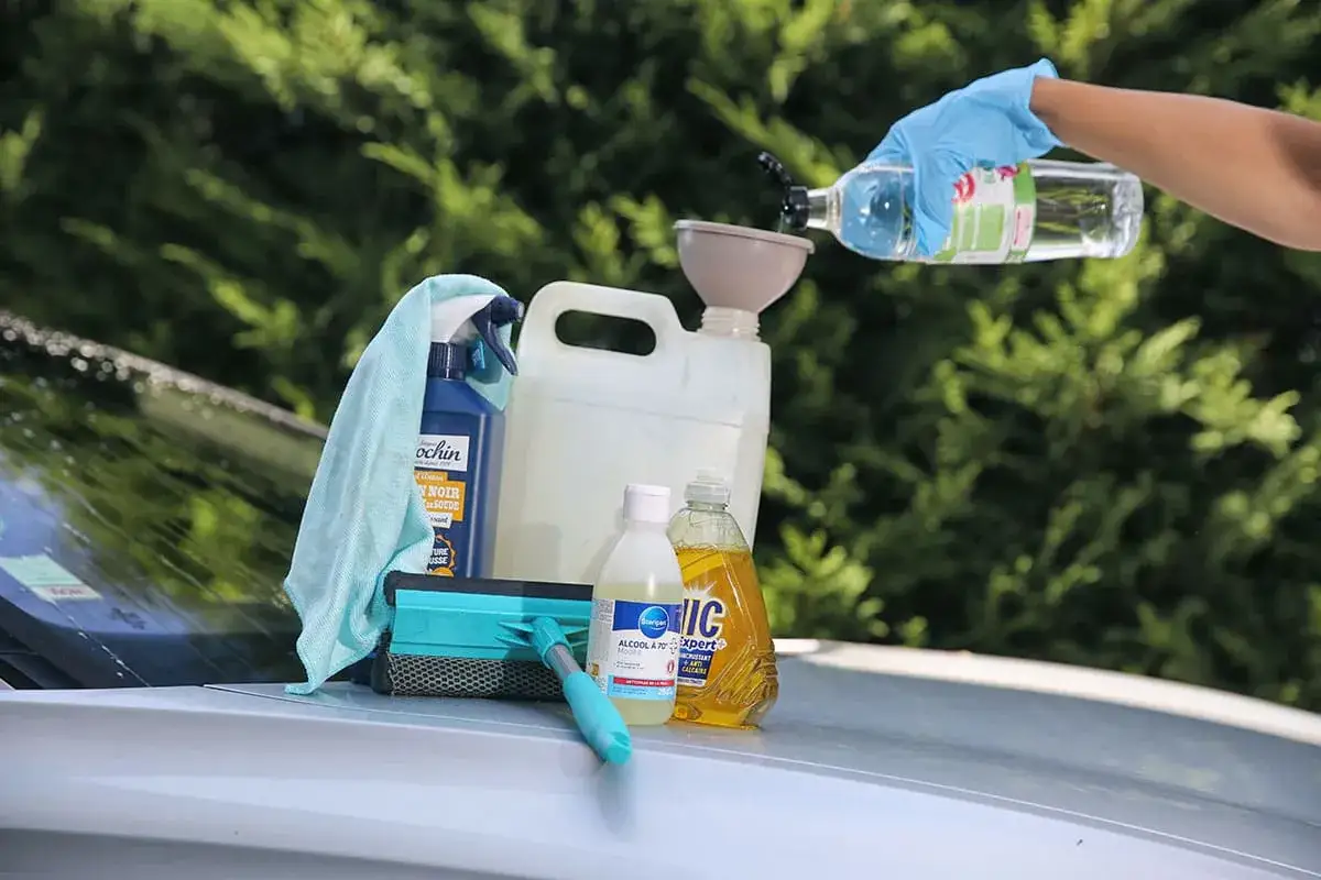 Préparation d'un lave glace maison : bidon, flacons d'alcool et liquide vaisselle, chiffon et raclette sur le capot d'une voiture.