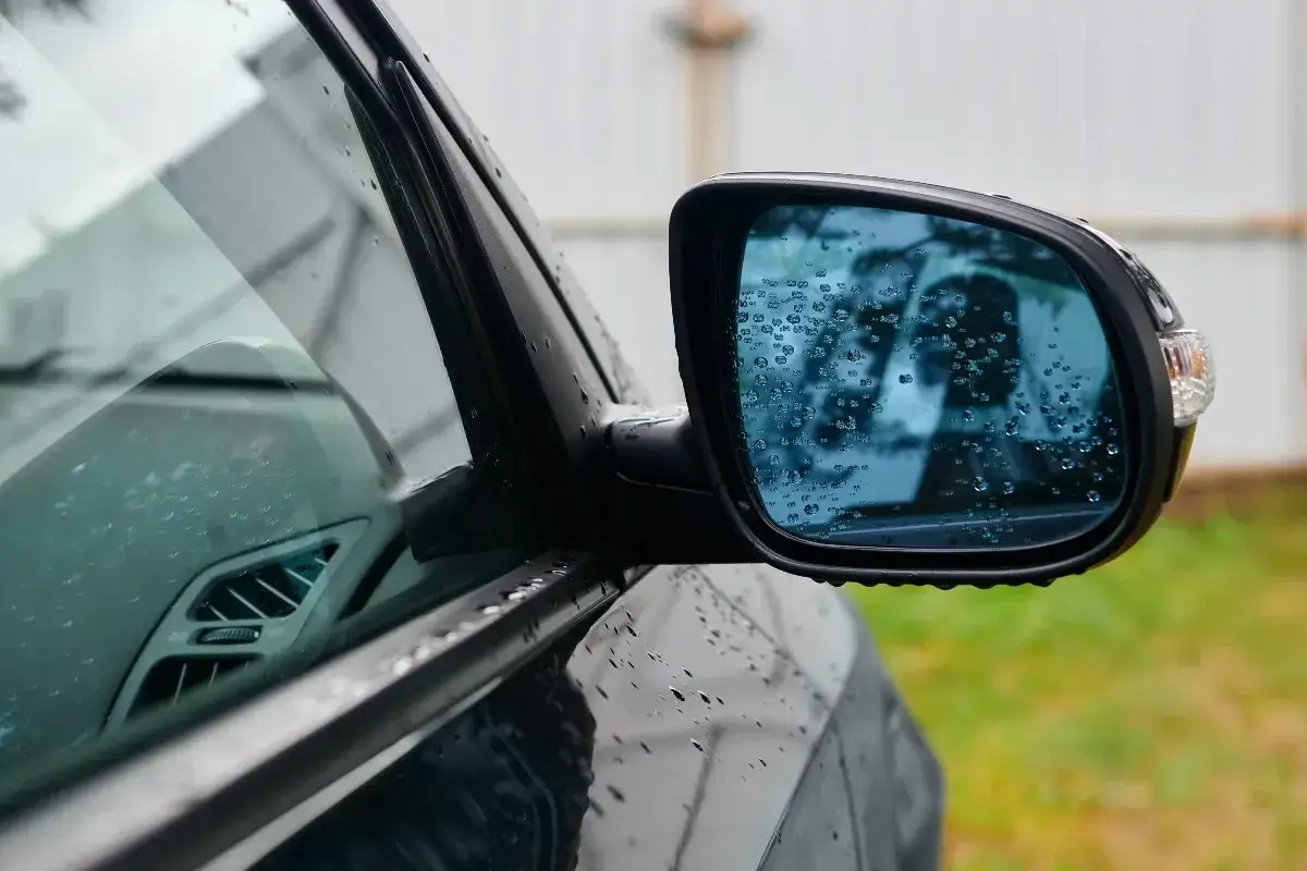 R&eacute;troviseur droit obligatoire depuis quand ? La pluie ruisselle sur le miroir bleu d'une voiture, refl&eacute;tant un paysage flou.